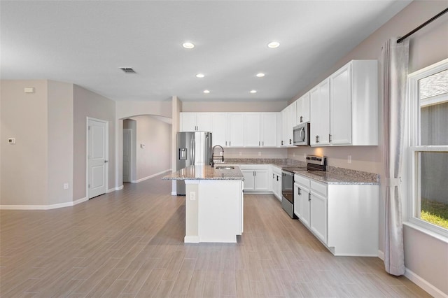 kitchen featuring light stone countertops, a kitchen island with sink, arched walkways, stainless steel appliances, and baseboards