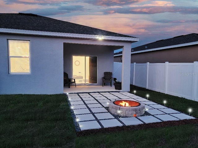 rear view of property featuring a shingled roof, a patio area, fence, a fire pit, and stucco siding