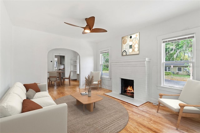 living area with a brick fireplace, arched walkways, ceiling fan, and wood finished floors