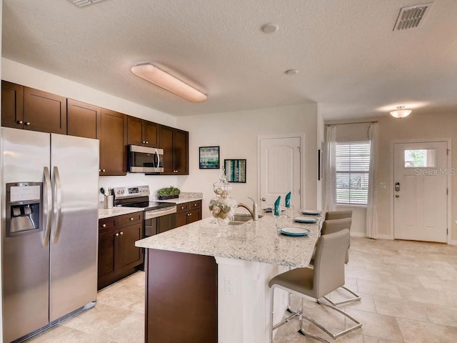 1550 Dalehurst Ln, Kissimmee, FL 34744 - kitchen with visible vents, dark brown cabinetry, a center island with sink, light stone countertops, and stainless steel appliances