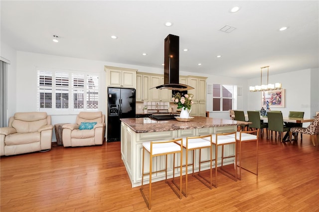 kitchen with black fridge with ice dispenser, a kitchen bar, cream cabinetry, dark stone counters, and island range hood