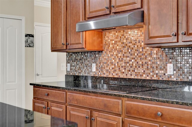 kitchen featuring tasteful backsplash, dark stone countertops, brown cabinetry, black electric cooktop, and under cabinet range hood