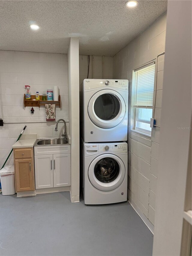 laundry area featuring stacked washing maching and dryer, a textured ceiling, a sink, and concrete block wall