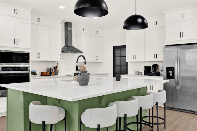 kitchen with an island with sink, tasteful backsplash, wall chimney range hood, white cabinetry, and stainless steel appliances