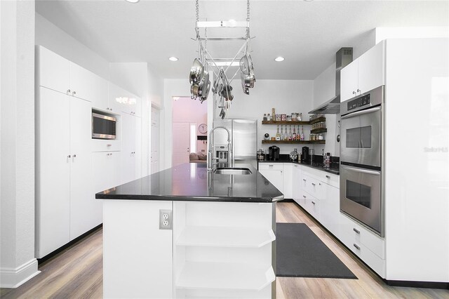 kitchen with wall chimney range hood, open shelves, dark countertops, appliances with stainless steel finishes, and a sink