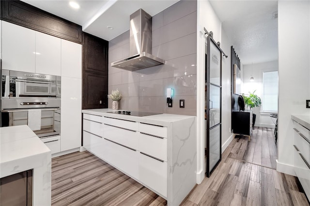 kitchen featuring wall chimney range hood, black electric stovetop, a barn door, light wood-style floors, and stainless steel oven
