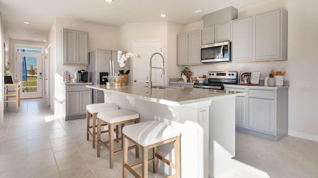 kitchen with a sink, an island with sink, stainless steel appliances, gray cabinets, and a breakfast bar area