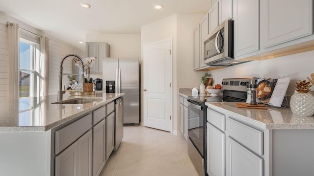 kitchen with a sink, an island with sink, gray cabinetry, stainless steel appliances, and light tile patterned flooring