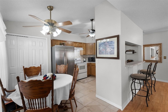 kitchen with brown cabinetry, electric range, a textured ceiling, freestanding refrigerator, and under cabinet range hood