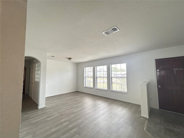 unfurnished living room with wood finished floors, visible vents, arched walkways, baseboards, and a textured ceiling