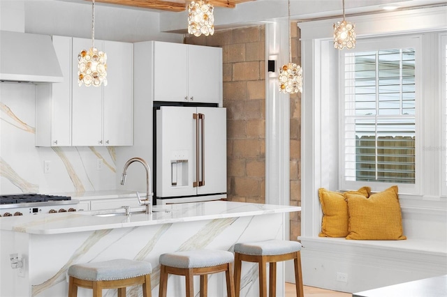 kitchen with high end fridge, wall chimney range hood, a sink, white cabinetry, and pendant lighting