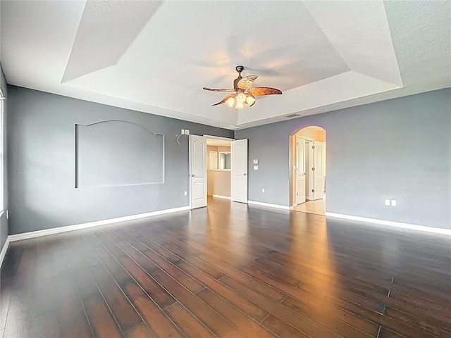 unfurnished room featuring a tray ceiling, arched walkways, baseboards, wood finished floors, and ceiling fan