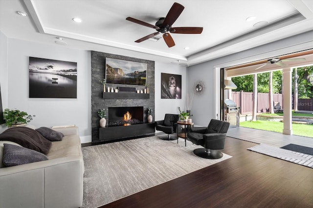 living area featuring recessed lighting, a tray ceiling, wood finished floors, a stone fireplace, and ceiling fan