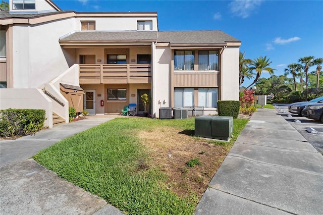 view of front facade featuring roof with shingles, stucco siding, a front yard, central AC unit, and a balcony