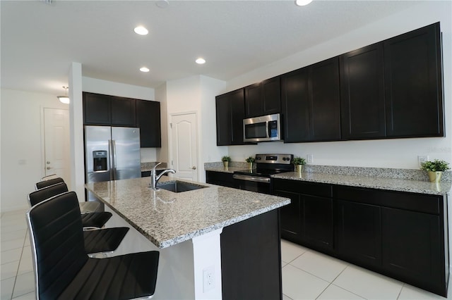kitchen featuring stainless steel appliances, a sink, dark cabinets, light stone countertops, and light tile patterned floors