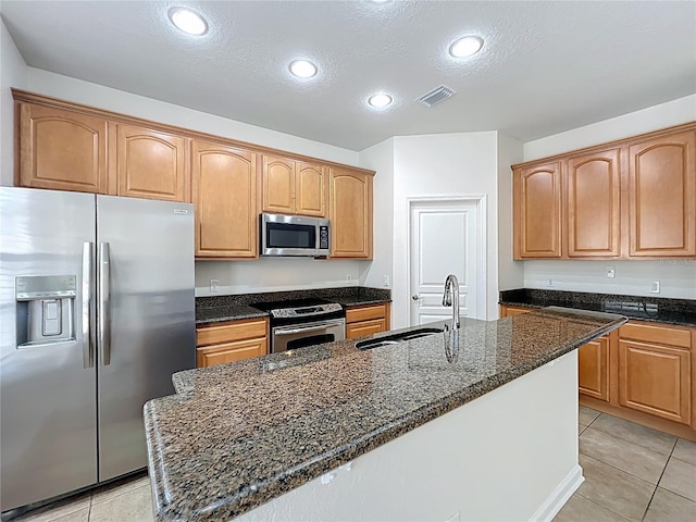 kitchen featuring an island with sink, stainless steel appliances, a sink, dark stone counters, and a textured ceiling