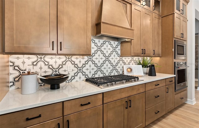 kitchen featuring light wood-style floors, backsplash, glass insert cabinets, custom range hood, and appliances with stainless steel finishes