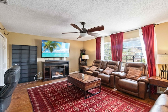 living room with wood finished floors, a glass covered fireplace, visible vents, a ceiling fan, and a textured ceiling