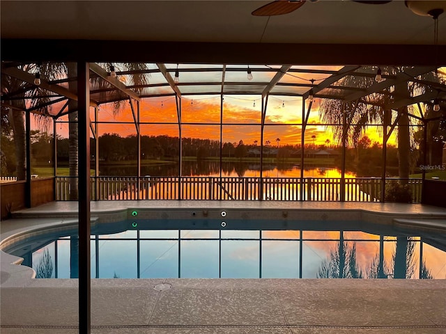 pool at dusk featuring a ceiling fan, a water view, glass enclosure, and an outdoor pool