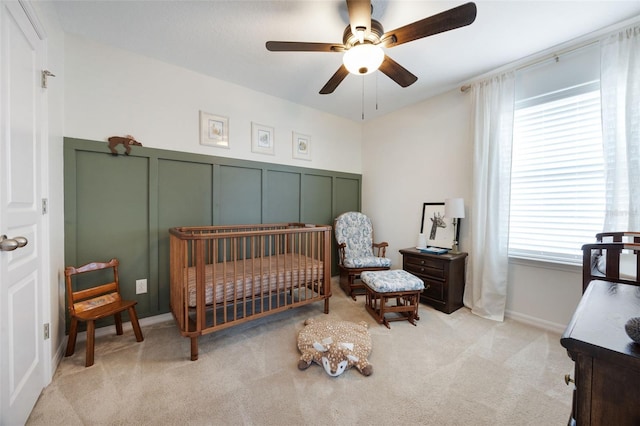 bedroom featuring a decorative wall, a crib, ceiling fan, a wainscoted wall, and carpet