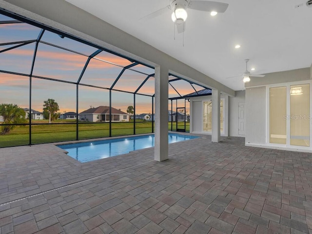 pool at dusk with glass enclosure, a lawn, an outdoor pool, a patio, and ceiling fan