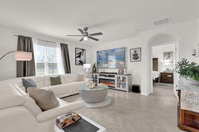 living room featuring light tile patterned flooring, a glass covered fireplace, visible vents, and ceiling fan