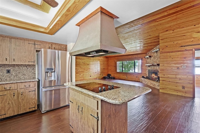 kitchen with dark wood-style flooring, wood walls, black electric cooktop, premium range hood, and stainless steel fridge