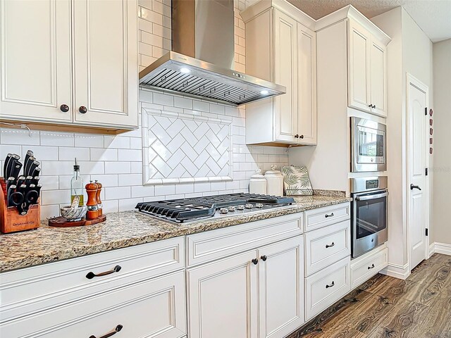 kitchen featuring wall chimney range hood, white cabinets, appliances with stainless steel finishes, light stone countertops, and dark wood-type flooring