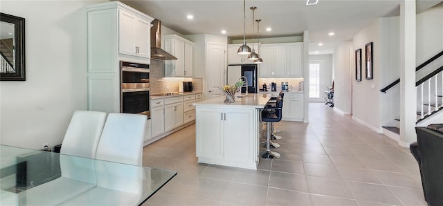 kitchen with wall chimney range hood, a breakfast bar area, backsplash, light countertops, and double oven