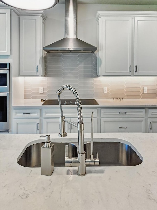 kitchen featuring stainless steel double oven, light stone counters, a sink, wall chimney exhaust hood, and decorative backsplash