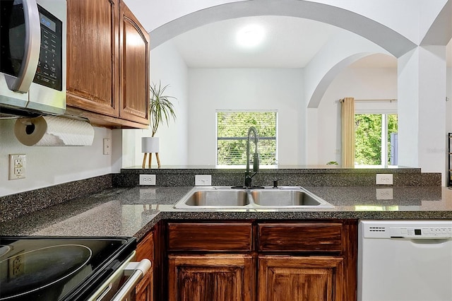 kitchen with dark stone counters, a sink, stainless steel appliances, brown cabinetry, and arched walkways