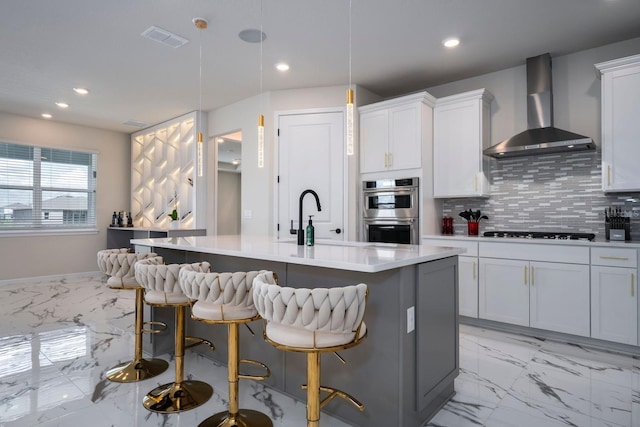 kitchen featuring a kitchen island with sink, wall chimney range hood, marble finish floor, double oven, and visible vents