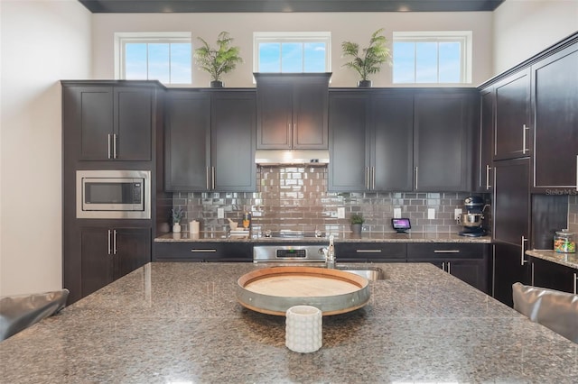 kitchen with stone counters, tasteful backsplash, stainless steel microwave, and under cabinet range hood
