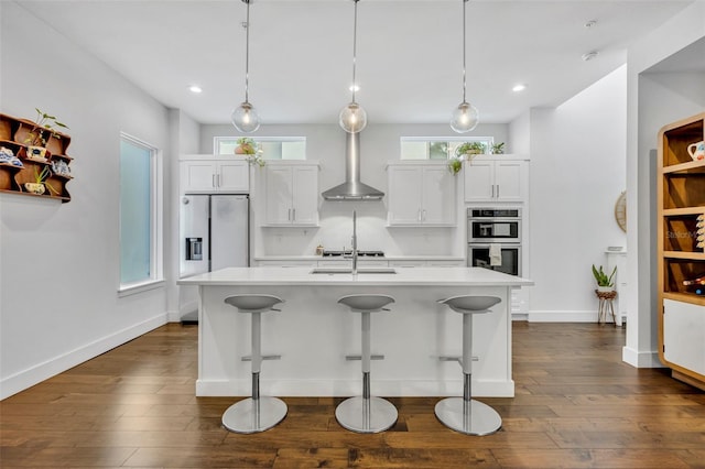 kitchen featuring light countertops, a center island with sink, dark wood-type flooring, wall chimney range hood, and appliances with stainless steel finishes