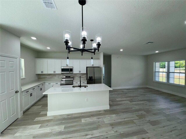 kitchen with a kitchen island with sink, appliances with stainless steel finishes, a sink, light wood finished floors, and an inviting chandelier