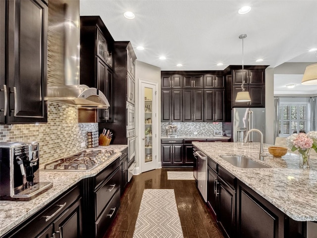 kitchen featuring wall chimney range hood, a sink, dark wood finished floors, an island with sink, and stainless steel appliances
