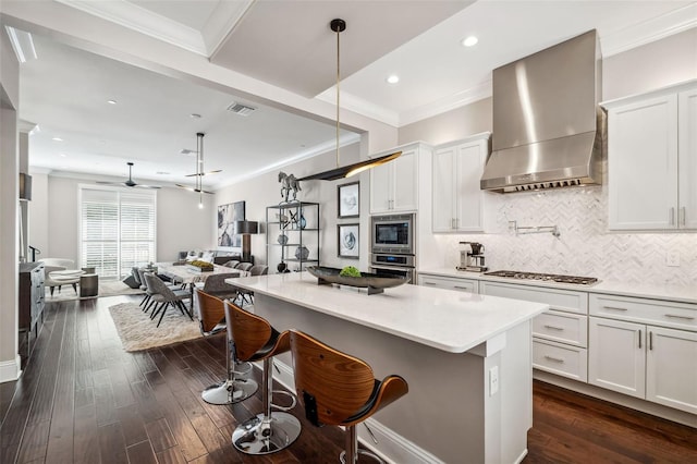 kitchen featuring wall chimney range hood, appliances with stainless steel finishes, tasteful backsplash, open floor plan, and ornamental molding