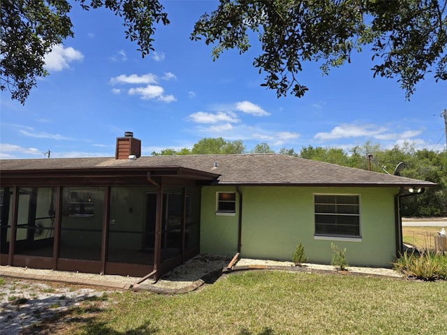 back of house featuring a shingled roof, stucco siding, a sunroom, a chimney, and a lawn