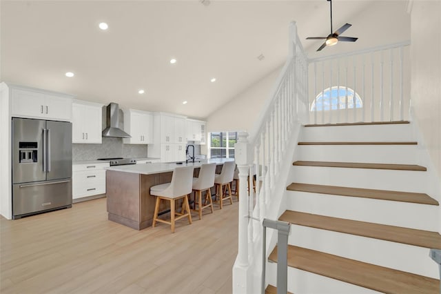 kitchen with wall chimney range hood, backsplash, a center island with sink, stainless steel fridge with ice dispenser, and white cabinetry