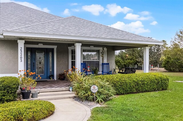 entrance to property featuring a porch, roof with shingles, stucco siding, and a yard