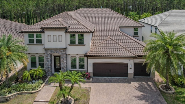 mediterranean / spanish-style home with stucco siding, an attached garage, a tiled roof, decorative driveway, and a view of trees