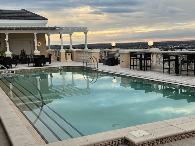 pool at dusk with an outdoor kitchen, outdoor dry bar, a pergola, and a patio