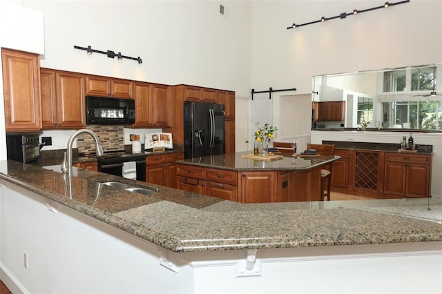 kitchen featuring brown cabinets, a sink, dark stone counters, a towering ceiling, and black appliances