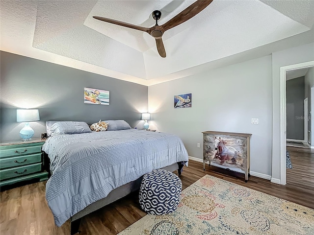 bedroom featuring a textured ceiling, a ceiling fan, baseboards, wood finished floors, and a tray ceiling