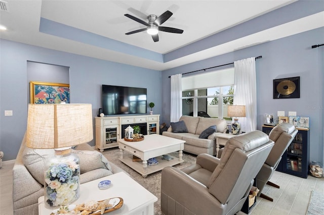 living area with ceiling fan, visible vents, light wood-type flooring, baseboards, and a tray ceiling