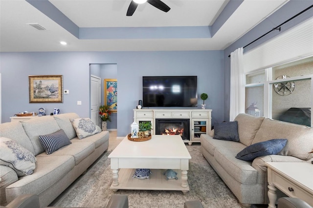 living area with a glass covered fireplace, a ceiling fan, recessed lighting, a tray ceiling, and visible vents