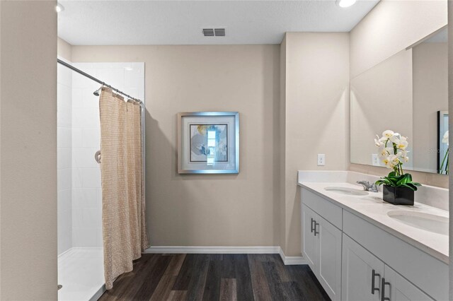 full bath featuring double vanity, a tile shower, wood finished floors, a sink, and baseboards