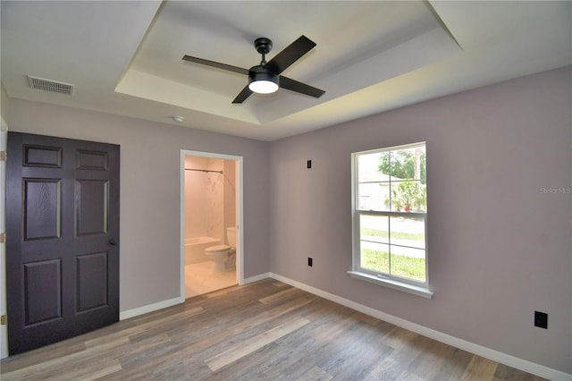 unfurnished bedroom with a tray ceiling, wood finished floors, baseboards, ensuite bathroom, and a ceiling fan