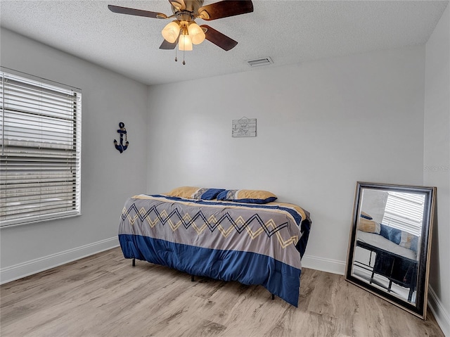 bedroom with ceiling fan, a textured ceiling, baseboards, wood finished floors, and visible vents