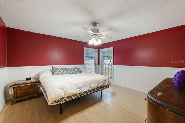 bedroom featuring a textured ceiling, light wood-style flooring, wainscoting, and ceiling fan
